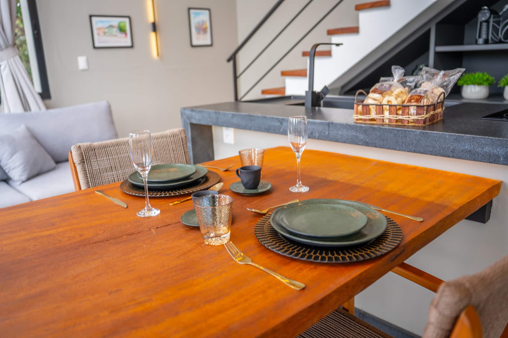 Mesa posta com pratos de cerâmica verde, taças e cesta de pães sobre a bancada ao fundo