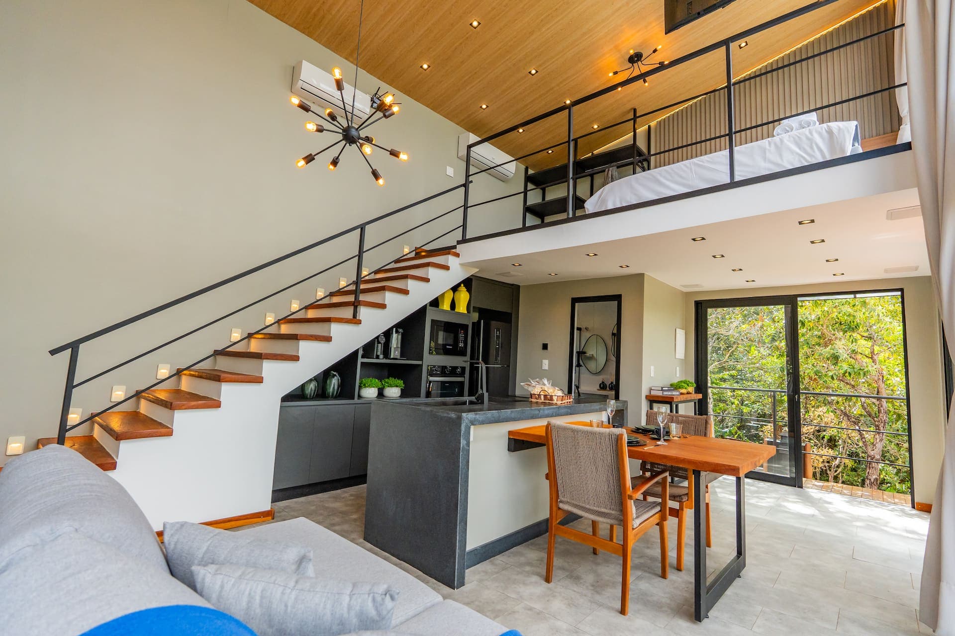 Escada de madeira sob o mezanino do quarto, com cozinha aberta e vista para a mata