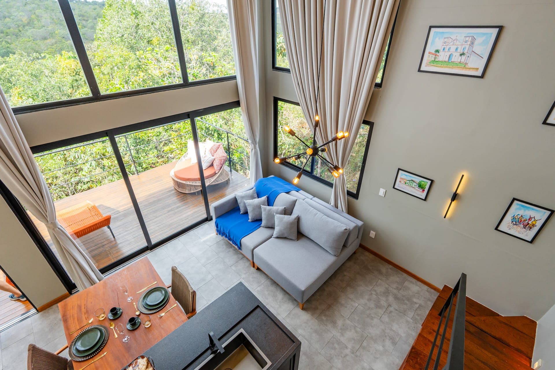 Vista aérea da sala com sofá cinza e manta azul, mesa de jantar posta e deck com espreguiçadeira laranja ao fundo